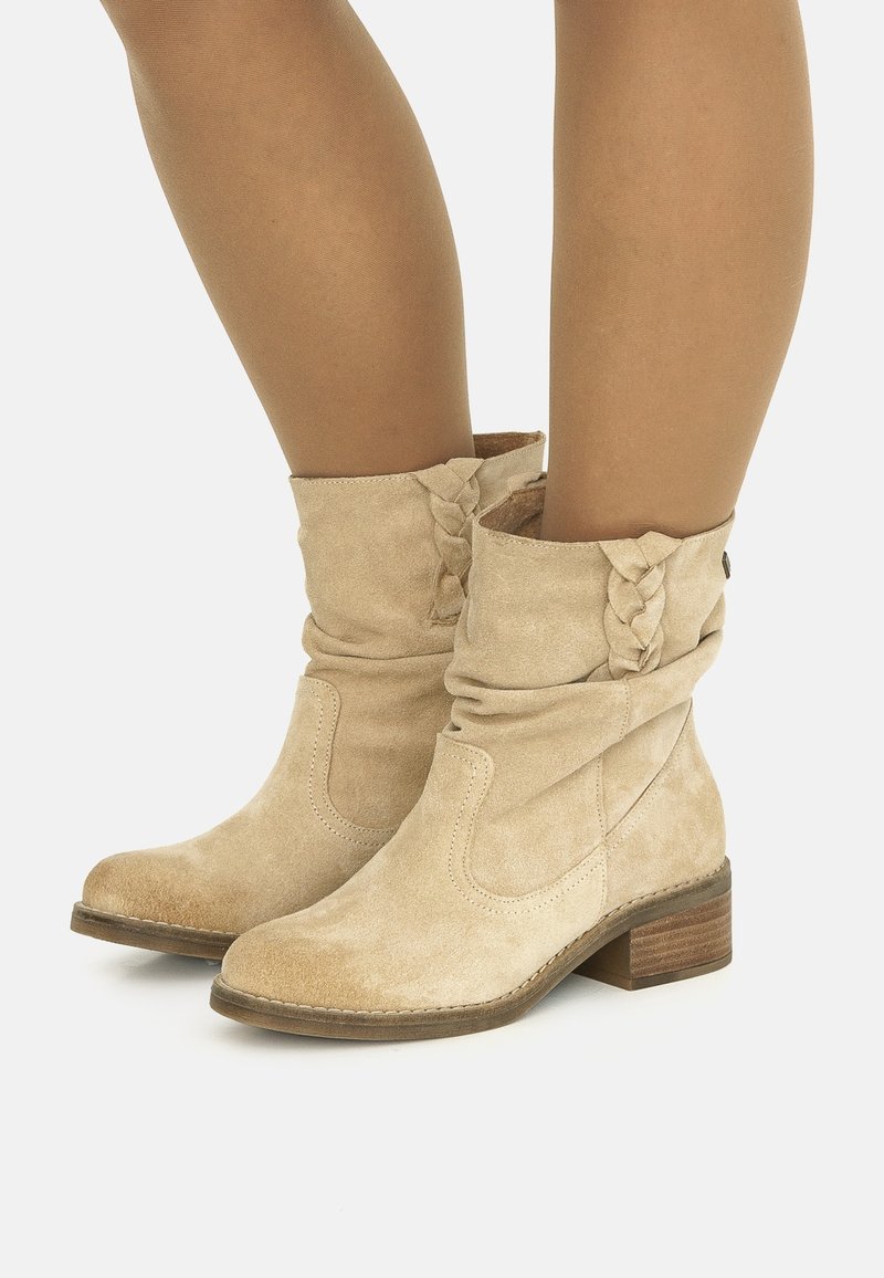 Bottes mi-mollet en daim beige avec un détail tressé et des talons en bois empilés bas, portées avec des collants beiges transparents sur fond blanc.