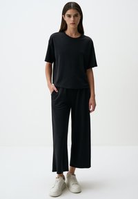 Zwarte katoenen t-shirt met korte mouwen, gecombineerd met bijpassende wijd uitlopende broek. Model staat in witte sneakers tegen een neutrale achtergrond.