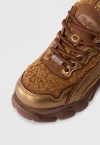 Καφέ αθλητικά παπούτσια με αφράτη υφή, χρυσές λεπτομέρειες, χοντρή σόλα και καφέ κορδόνια, με ετικέτα μάρκας στο πλάι.