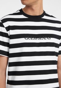 Černobíle pruhované tričko s kulatým výstřihem, které na přední straně obsahuje vyšívané logo "GUESS JEANS". Krátké rukávy a bavlněný materiál.