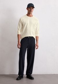 Helles gelbes Langarmshirt, marinefarbene Hose, schwarzer Gürtel und schwarze Schuhe, die von einem Mann getragen werden. Einfarbiges Design mit lockerer Passform.