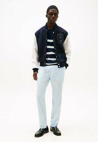 Marineblau und weiße Collegejacke mit H-Logo, gestreiftes Marineblau- und Weißhemd darunter, hellblaue Hose und schwarze Schuhe. Glatte Texturen.
