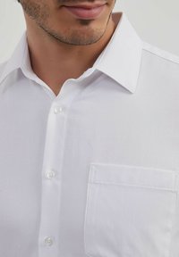 Chemise blanche à boutons, en tissu texturé, avec un col pointu et une poche avant unique. Équipée de boutons nacrés et de coutures soignées.