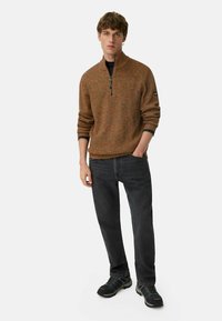 Ung mand iført en brun sweater med halv lynlås, sort skjorte underneath, sorte jeans og sorte sneakers, stående med den ene hånd i lommen.