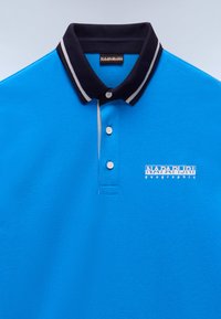 Blå polo med marinblå krage, vita ränder, tre knappar och en logotyp på vänster bröst. Tillverkad av strukturerad bomullstyg.