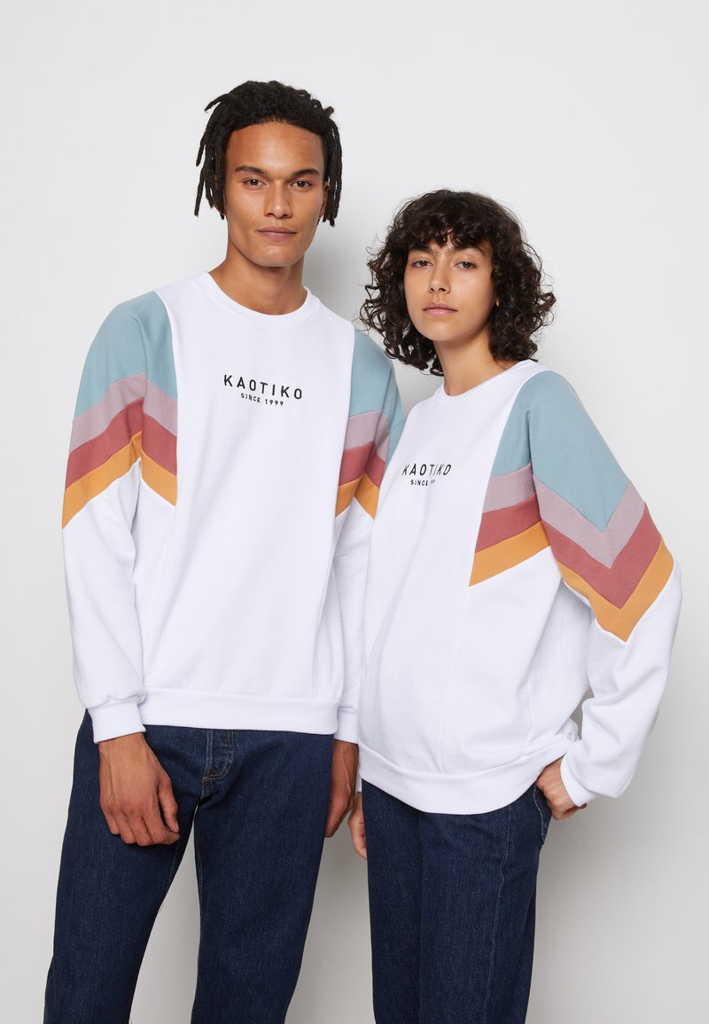 Hvide sweatshirts med farverige chevron mønstre i pastelfarverne blå, pink, sennep og terracotta, med en central logo tekst.