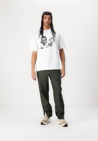 White cotton t-shirt with black graphic print, paired with green loose-fit pants and beige sneakers with dark accents.