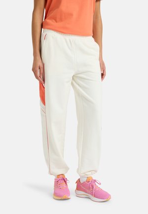 Witte joggers met een oranje paneel aan de zijkant, elastische tailleband en rode accenten. Gepaard met roze sportschoenen met witte zolen.