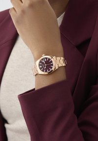 Reloj de pulsera en oro rosa con esfera burdeos, números romanos y diseño de pulsera eslabonada. Lleva puesto en una muñeca con una manga granate.