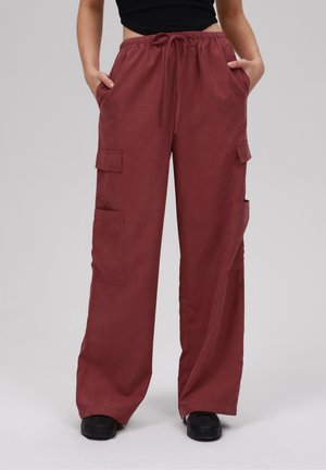 Femme portant un pantalon cargo ample bordeaux avec poches latérales et baskets noires, mains dans les poches, sur un fond clair uni.