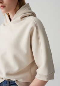 Crème-kleurige hoodie van zachte stof, met een capuchon en korte mouwen met ribgebreide manchetten. Naaddetails benadrukken het ontwerp.