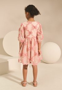 Vestido floral rosa con mangas abullonadas, falda de volantes, tejido texturizado y adornos decorativos. Sandalias complementan el atuendo.