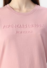 Vaaleanpunainen puuvillapaita, jossa on kohokirjaimin painettu teksti "PEPE JEANS LONDON" ja "WEST ELEVEN" edessä, pyöreä pääntie ja pehmeä rakenne.
