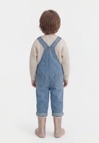 Kind mit kurzen braunen Haaren, das rückwärts schaut, trägt einen beigefarbenen Zopfmuster-Pullover, blaue Denim-Latzhose und rosa gemusterte Stiefel vor einfachem Hintergrund.