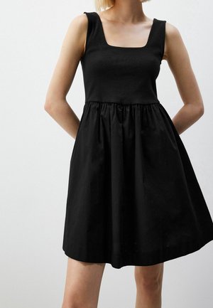 Woman wearing a sleeveless black dress with a square neckline and gathered skirt, standing with hands behind her back.