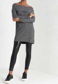 Graues, schulterfreies Strickkleid mit langen Ärmeln, kombiniert mit glänzenden schwarzen Leggings und schwarzen Sneakers mit Schnallenakzenten.