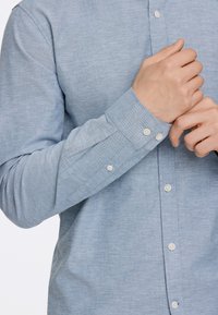 Camisa de botones azul claro con tela texturada, de manga larga y botones blancos. Los puños presentan un diseño simple con detalle de costura.