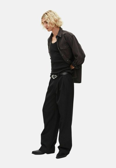 Νεαρός με ξανθά σγουρά μαλλιά, φορώντας ένα σκούρο ριγέ πουκάμισο πάνω από μια μαύρη αμάνικη μπλούζα, φαρδιά μαύρα παντελόνια και μαύρα παπούτσια, στέκεται πλαγιαστά.