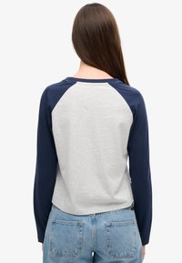 Vrouw met lang bruin haar draagt een grijze shirt met donkerblauwe lange mouwen en lichtblauwe spijkerbroek, gezien van achteren.