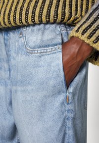 Lysblå denimjeans med en frontlomme, fremhævet af orange syninger. Stoffet har en subtil tekstur, som er kombineret med en tekstureret gul og sort sweater.
