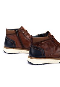 Zapatos de cuero marrón con un acabado texturizado, que incluyen una cremallera lateral, acentos en contraste en azul marino y una suela de goma de color claro.