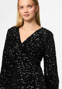 Vestido negro de lentejuelas con mangas largas y escote en V. La tela presenta una superficie texturizada con lentejuelas circulares brillantes.