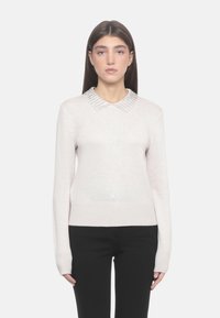 Suéter de un color beige claro con textura acanalada y mangas largas. Presenta un cuello decorativo con cuentas. Se lleva con pantalones negros.