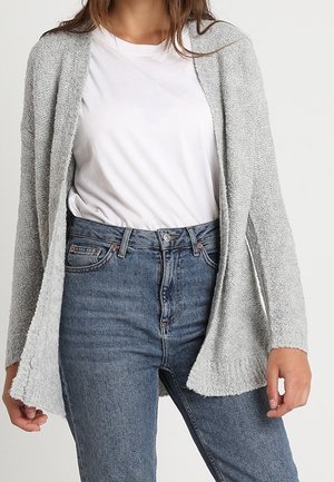 Mujer con un cárdigan texturizado gris claro sobre una camiseta blanca lisa metida en vaqueros azules de talle alto sobre un fondo neutro.