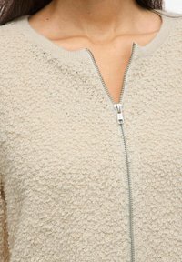 Beige krēmkrāsas zip-up džemperis ar strukturētu, mīkstu virsmu un ribota kakla izgriezumu. Centrā ir sudraba rāvējslēdzis. Mīksta auduma.