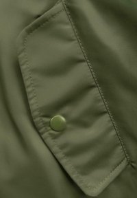 Alyvuogių žalia medžiaga su tekstūruotu paviršiumi. Turi didelę kišenę su atvartais, pritvirtintą žaliu apvaliu spaustuku. Matomos siūlių detalės.