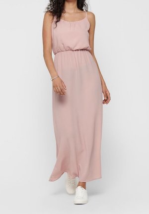 Femme portant une robe longue rose pâle sans manches avec une taille élastique, associée à des baskets blanches, debout sur un fond clair uni.