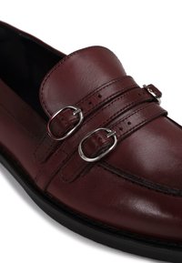 Mocassins de couro cor burgundy com design de dupla fivela em prateado, bico arredondado e costura texturizada na parte superior. Sola preta.