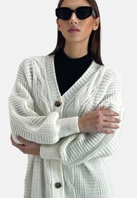 Weißer Strickcardigan mit strukturierten Mustern, übergroßen Ärmeln und V-Ausschnitt. Verfügt über einen einzelnen braunen Knopf und gerippte Bündchen.