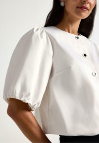 Hvid bluse med puffede ærmer, rund halsudskæring og sorte knapdetaljer. Glat stof med let glans.