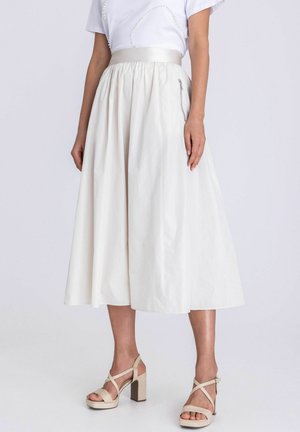 Frau trägt einen weißen Midirock mit geraffter Taille und seitlichem Reißverschluss, kombiniert mit beigen Sandalen mit hohen Absätzen und Knöchelriemen.