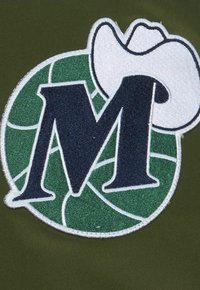 Insigne brodé avec une lettre "M" marine surmontée d'un chapeau de cowboy blanc, entourée d'un design circulaire vert sur un fond olive.