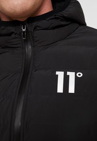 Chaqueta acolchada negra con cierre de cremallera, que cuenta con capucha y un logo impreso en blanco "11°" en el frente. Textura suave, diseño minimalista.