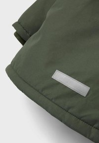 Giacca verde imbottita con una texture liscia, dotata di un'etichetta rettangolare argentata sull'orlo. Il design ha un aspetto strutturato e moderno.