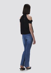 Top nero off-shoulder con maniche a soffietto, abbinato a jeans a zampa d'elefante blu. Modella vista di spalle, con tacchi neri.