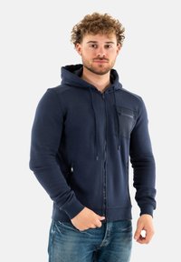 Felpa con zip di colore blu navy realizzata in tessuto morbido, presenta una tasca frontale con zip, cappuccio regolabile concordino e polsini a coste. Indossata con jeans blu.