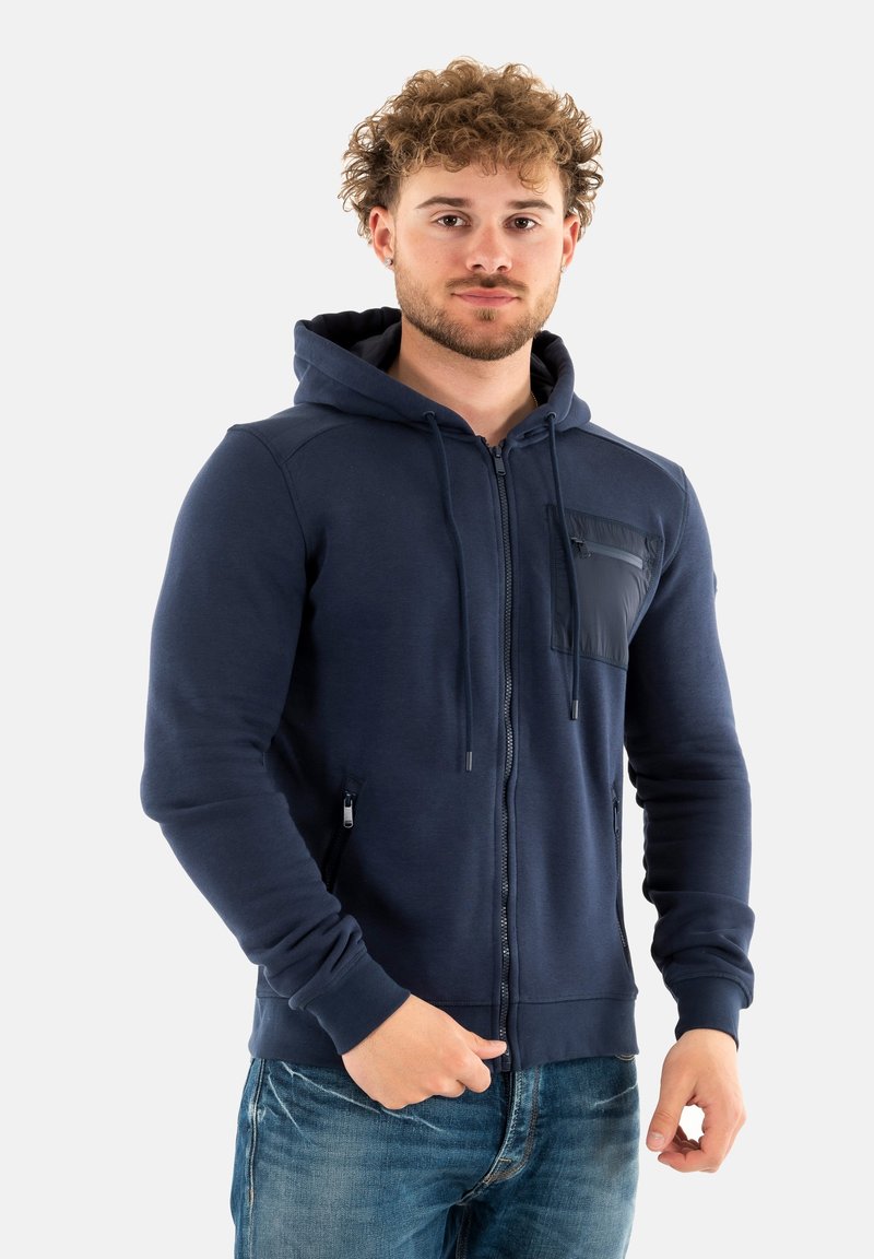 Felpa con zip di colore blu navy realizzata in tessuto morbido, presenta una tasca frontale con zip, cappuccio regolabile concordino e polsini a coste. Indossata con jeans blu.