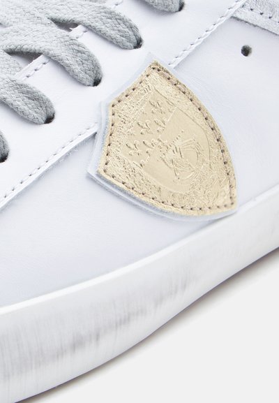 Άσπρο αθ λητικό παπούτσι με λείο δέρμα στο πάνω μέρος, γκρι κορδόνια και υφασμένο μπεζ έμβλημα στο πλάι, με περίτεχνη λεπτομέρεια.