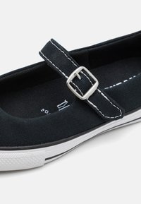 Zapatilla de lona negra con costuras blancas en contraste, una correa con hebilla y una suela de goma que presenta una puntera blanca texturizada y rayas negras.
