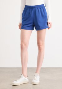 Calções desportivos azuis com um cós elástico, tecido suave e bolsos laterais. Usados com ténis brancos com uma sola texturizada.