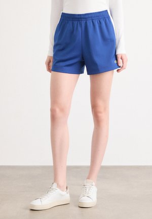 Blue athletic shorts with an elastic waistband, smooth fabric, and side pockets. Worn with white sneakers featuring a textured sole.