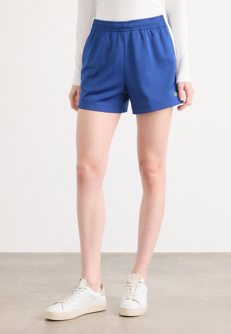 Calções desportivos azuis com um cós elástico, tecido suave e bolsos laterais. Usados com ténis brancos com uma sola texturizada.