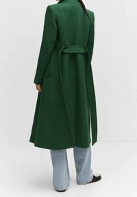 Long manteau vert en tissu texturé, doté d'une ceinture nouée à la taille et d'une fente au dos, associé à un jean évasé bleu clair et à des chaussures noires.