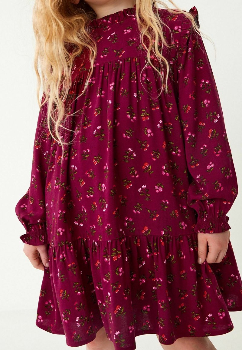 Enfant portant une robe à manches longues, de couleur rose foncé avec un motif floral, un col à volants et une jupe froncée, debout contre un arrière-plan clair.