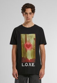 Melns kokvilnas T-krekls ar lielu sirds grafiku meža ainavā, kurā vārdam "L.O.V.E." ir uzrakstīts treknrakstā baltām burtiem.