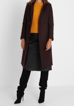 Femme portant un long manteau marron foncé, un pull jaune moutarde, une jupe en cuir noire et des bottes noires à talons hauts, debout devant un fond blanc.
